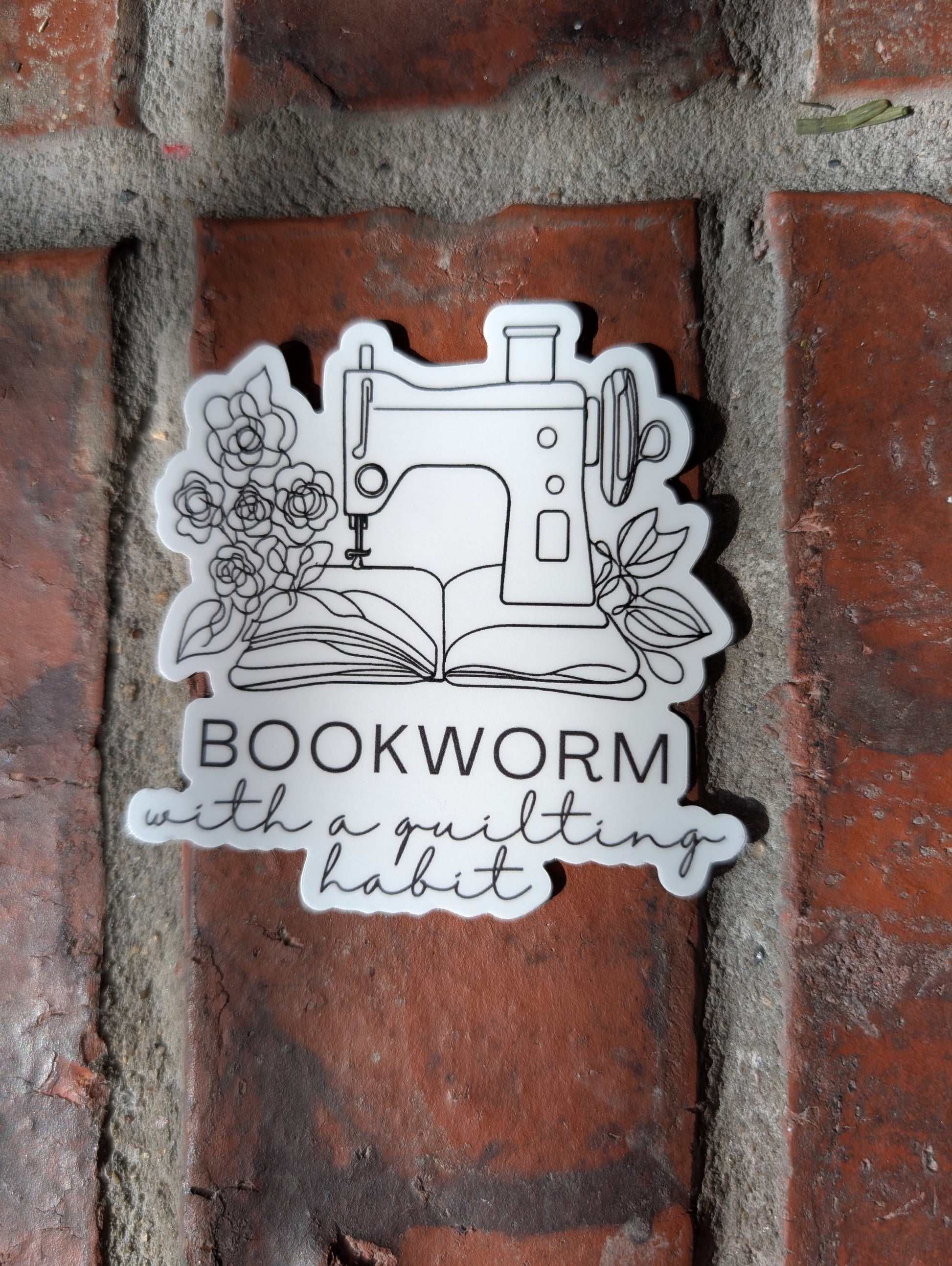 Sticker with sewing machine design and 'Bookworm with a quilt' text on a brick wall.